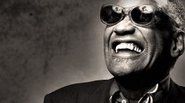 Ray Charles smiling in a collared shirt and sports jacket wearing his trademark sunglasses.