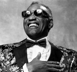Ray Charles in a floral print suit with a bow tie, sunglasses, and a hand over his heart